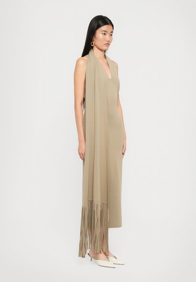 Woman with long black hair wearing a sleeveless beige dress with a deep V-neck and long fringe details at the hem, paired with white heels.