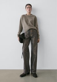 Pull marron clair avec des poignets côtelés, associé à un jean bronze foncé à coupe droite et léger éclat. Tient un sac à main foncé.