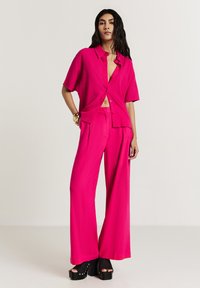 Camicetta rosa brillante a maniche corte abbinata a pantaloni larghi coordinati. Tessuto testurizzato, vestibilità comoda, con bottoni e design corto. Sandali con tacco alto.