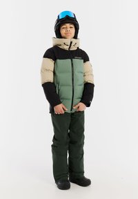 Jugend-Puffjacke in Grün, Schwarz und Beige, mit hohem Kragen, Frontreißverschluss und elastischen Bündchen. Getragen mit dunkelgrünen Hosen und schwarzen Stiefeln.