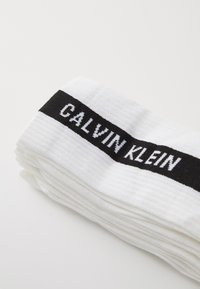 Hvide ribbede sokker med et sort bånd, der har "CALVIN KLEIN" i hvid skrift. Stoffet ser blødt ud med en glat tekstur.