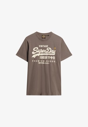 T-shirt di cotone marrone con maniche corte, che presenta testi bianchi e dorati che leggono "VINTAGE SuperDry PREMIUM GOODS" e caratteri giapponesi.