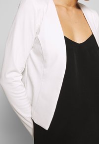 Vit, figursydd kavaj med en slät textur, öppen front och l ånga ärmar, bärs över en svart topp och visar en ren, modern design.