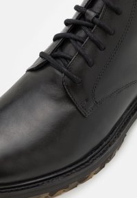 Bota de cuero negro con textura suave, cordones resistentes y suela rugosa con un patrón contrastante. Detalles de costura visibles.