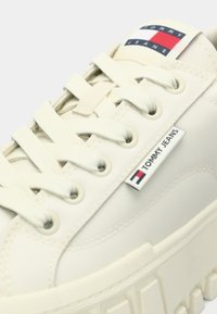 Δερμάτινο αθλητικό παπούτσι σε εκρού απόχρωση με επίπεδα κορδόνια, λαστιχένια σόλα και ετικέτα με το λογότυπο που φέρει κόκκινες, λευκές και μπλε λεπτομέρειες στη γλώσσα.