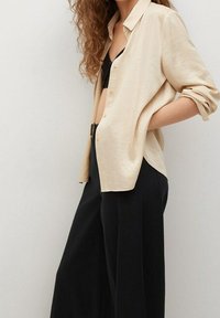 Béžová, lehká košile na knoflíky s vyhrnutými rukávy, v kombinaci s černými širokými kalhotami. Hladká textura, volný střih, neformální design.