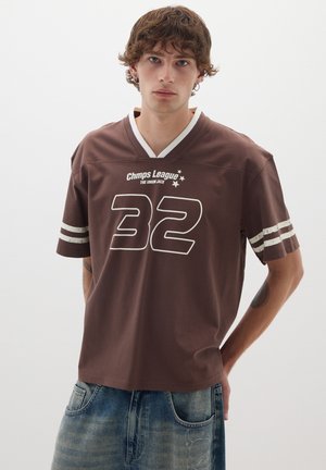 Jersey marrón de manga corta con acentos blancos, con cuello en V, gráfico grande "32" y texto "Chmps League". Combinado con shorts de mezclilla azules.
