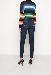 Jeans ajustados de mezclilla en un color azul oscuro, combinados con un suéter a rayas multicolor que presenta una textura acanalada y colores de mangas contrastantes. Tacones altos negros visibles.
