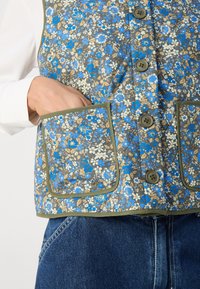 Ανθοστολισμένο γιλέκο με μπλε και καφέ σχέδιο σε γκρι φόντο, πέντε κουμπιά και ελιά πράσινη διακόσμηση γύρω από τις τσέπες.