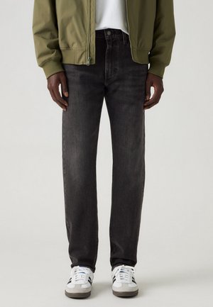 Homme portant un jean droit noir, des baskets blanches avec des bandes noires, une chemise blanche et une veste zippée vert olive, debout devant un fond uni.