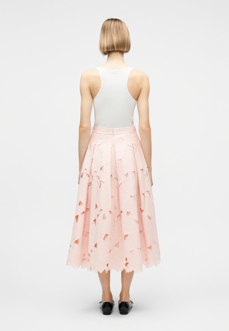 Donna con capelli biondi corti che indossa un top bianco senza maniche e una gonna midi rosa con motivi floreali traforati, vista da dietro su uno sfondo bianco.