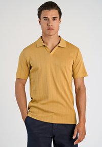 Kortærmet polo-shirt i sennep med V-hals, der har et struktureret vertikalt mønster og blødt strikket stof. Sammen med mørke bukser.