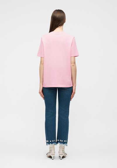 Γυναίκα που φοράει ανοιχτό ροζ κοντομάνικο πουκάμισο, μπλε κοντά τζιν με floral διακοσμήσεις και λευκές πέδιλες με τακούνι, στέκεται προς τα πίσω.