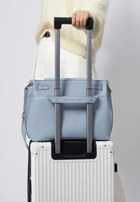 Ljusblå läderhandväska placerad på en vit resväska med texturerad yta, utrustad med två grå teleskophandtag. Accenterna inkluderar guldfärgad metall.