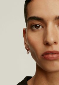 Pendiente de aro dorado con un colgante de cristal transparente, que presenta una textura suave y un acabado pulido, llevado en la oreja de una persona.
