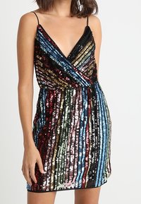 Frau, die ein ärmelloses, V-ausschnitt Mini-Kleid mit vertikalen, mehrfarbigen Paillettenstreifen und dünnen schwarzen Schulterträgern trägt.