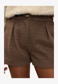 Pantalones cortos de lana marrón con textura suave, con pliegues frontales y cierre de botón. Combinados con un suéter de punto de color claro.