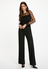 Zwarte transparante blouse met polkadots en kanten details, gecombineerd met hoge zwarte wijde broek en puntige zwarte hakken.