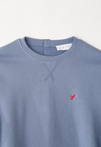 Felpa blu con scollatura rotonda e polsini a costine, caratterizzata da un piccolo logo di un uccellino ricamato di colore rosso sul petto. Tessuto morbido.