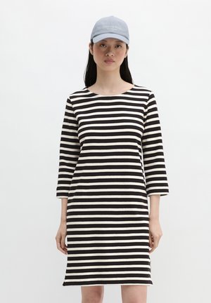 Vrouw die een zwart-wit horizontaal gestreepte jurk draagt met driekwart mouwen en een lichtblauw baseballpetje tegen een effen witte achtergrond.