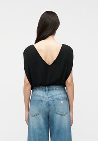 Γυναίκα με μακριά σκουρόχρωμα μαλλιά που φοράει μαύρο τοπ με βαθύ V πίσω και ψηλόμεσα ανοιχτό μπλε παντελόνια με ευρύ πόδι σε απλό φόντο.