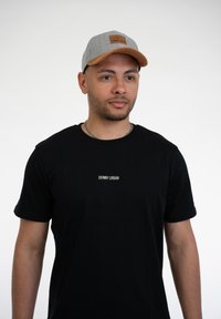 Harmaa lippalakki, jossa on ruskea nahkalappu, ja musta lyhythihainen t-paita, jossa on valkoisella tekstillä "JOHNNY URBAN". Neutraali tausta.