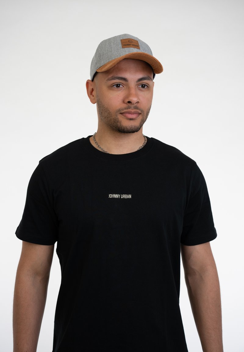 Harmaa lippalakki, jossa on ruskea nahkalappu, ja musta lyhythihainen t-paita, jossa on valkoisella tekstillä "JOHNNY URBAN". Neutraali tausta.