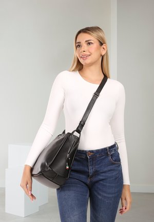 Woman wearing white long-sleeve top and blue jeans carries a black leather shoulder bag with an adjustable strap, standing indoors.