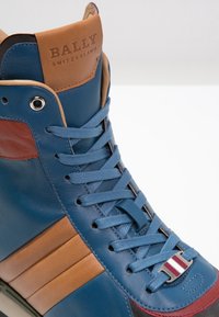Högsko-sneakers med blått läder, bruna accenter och blå snören. Anmärkningsvärda detaljer inkluderar en märkeslogotyp i tanfärgat läder och silverfärgade detaljer.