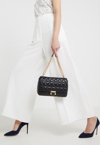 Černá prošívaná kabelka se zlatým řetězovým popruhem, obdélníkový tvar, se zlatým zapínáním. Kombinována s bílými kalhotami s širokými nohavicemi a modrými podpatky.