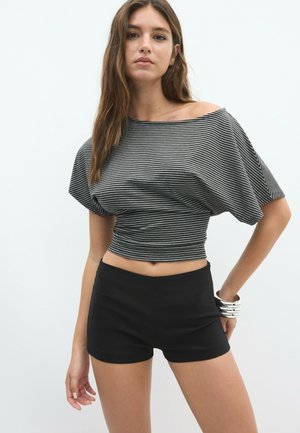 Kvinde iført en grå og sort striped off-shoulder crop top og sorte shorts, poserende med den ene hånd på hoften og sølv armbånd.