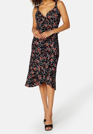Mujer con vestido midi negro con estampado floral en rojo y rosa, tirantes finos, escote en V y sandalias negras de tacón abierto, de pie con las manos juntas.