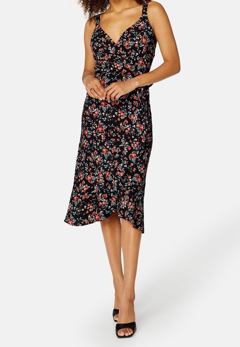 Femme portant une robe midi noire à imprimé floral rouge et rose, fines bretelles, encolure en V, et sandales à talons ouvertes noires, debout les mains jointes.