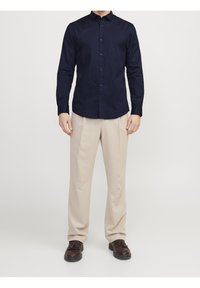 Camisa azul marino abotonada con mangas largas, combinada con pantalones beige claro y zapatos marrón oscuro. Tejido liso y diseño ajustado.