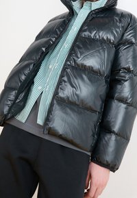 Chaqueta acolchada negra con textura brillante y acolchada y cuello alto, llevada sobre una camisa de botones a rayas verdes y blancas y pantalones oscuros.