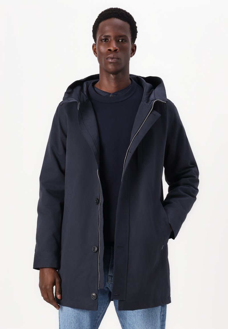 Granatowy wodoodporny płaszcz z kapturem, z zamkiem błyskawicznym, zatrzaskami i bocznymi kieszeniami. Gładka faktura i średnia długość.