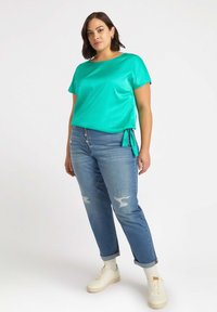 Turkoskortärmad topp med en knyt-detalj på sidan, tillsammans med ljusblå slitna jeans och vita sneakers. Slät textur.