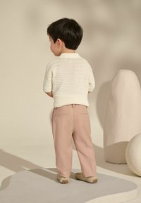 Weißes kurzärmliges Polohemd aus Strick, hellrosa Hosen, beigen Slippern, stehend mit dem Rücken zur Kamera in einem sanft beleuchteten, neutralen Raum.