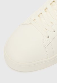 White leather sneaker featuring flat laces, smooth surface, rounded toe, and rubber sole with textured details.