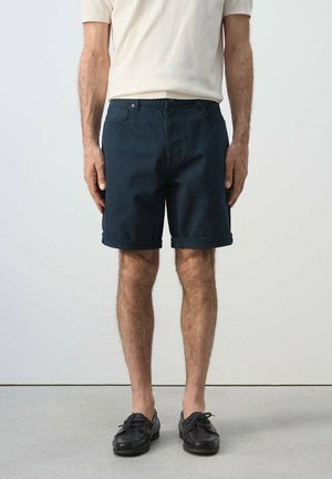 Man met donkerblauwe shorts met opgerolde zoom, zwarte loafers en een crème-kleurig overhemd met korte mouwen, staand tegen een effen lichte achtergrond.