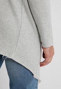 Hellgrauer Pullover mit geschwungener Saum und rohen Kanten, aus weichem Stoff. Detaillierte Ansicht von Arm und Hand auf Jeans.