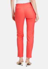 Korálové pánske nohavice so slim fit strihom, hladkou textúrou a zadnými vreckami, v kombinácii s metalickými vysokými sandálmi.