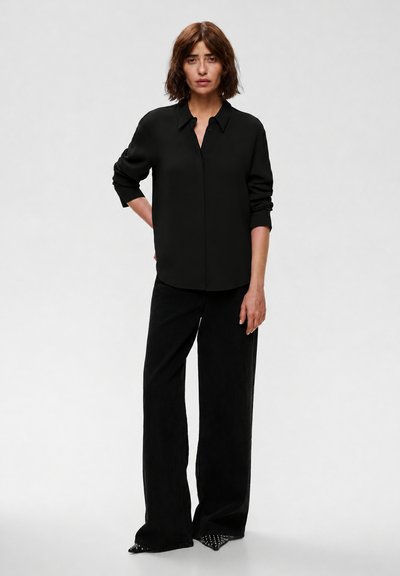 Femme debout devant un fond uni, portant une chemise noire à manches longues, un pantalon noir à jambes larges et des chaussures pointues cloutées.