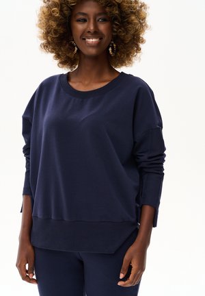 Uśmiechnięta kobieta o kręconych włosach nosi granatowy długi rękaw i spodnie, stoi na tle jednolitego białego tła.