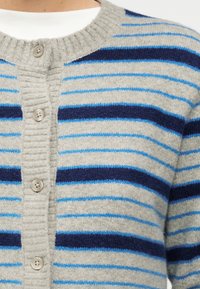 Κοντινή όψη γκρι ζακέτας με κουμπιά, με οριζόντιες μπλε και ναυτικές ρίγες, που φοριέται πάνω από ένα λευκό πουκάμισο.