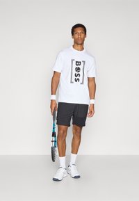 T-shirt blanche à manches courtes avec logo "BOSS" noir, assortie à un short noir. Le modèle tient une raquette de tennis et porte des sneakers blanches.