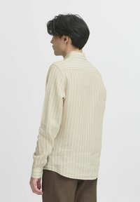 Beige langermet skjorte med tynne vertikale striper, strukturert stoff og høy krage. Klippet med rett fåll og knappedeler.