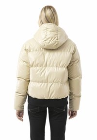 Beige Steppjacke mit glänzendem Finish, mit Kapuze, geripptem Saum und horizontalen Steppnähten. Schwarze Hose mit verzierten Gesäßtaschen.