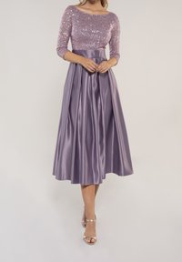 Ein lilafarbnes Midikleid mit einem pailettenverzierten Oberteil, Dreiviertelärmeln und einem glatten, plissierten Satinrock. Goldene Absatzsandalen.