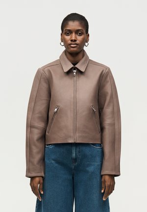 Femme portant une veste en cuir taupe avec des poches zippées, debout droite, associée à un jean denim bleu ample sur un fond uni.
